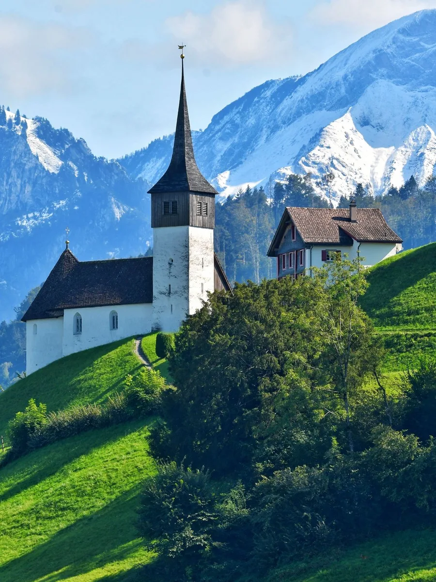 Weiße Kirche mit Spitzturm auf einer Wiese vor verschneiten Bergen